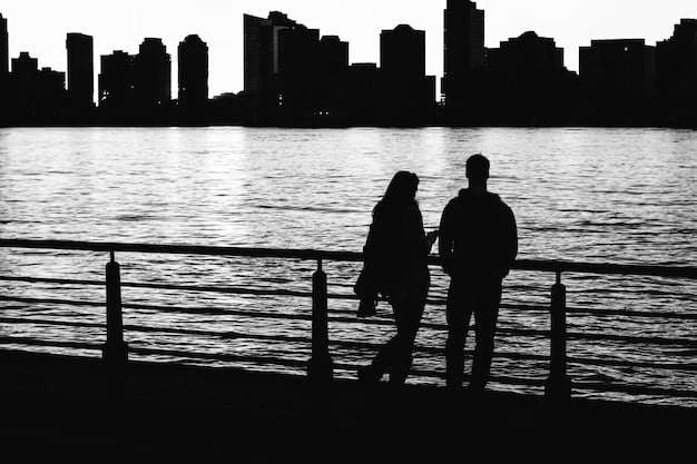 A couple holding hands, silhouetted against a sunset skyline. They are standing on opposite sides of a street or path, emphasizing the physical distance but also the connection they maintain. The image conveys a sense of longing and anticipation.