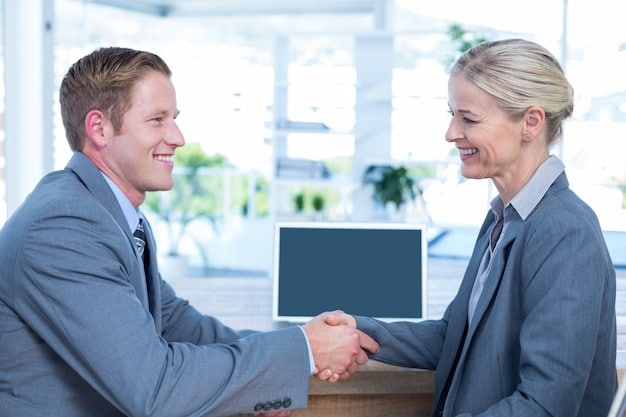 Two business people shaking hands across a table, with a laptop showing a graph. One is a person of color, the other is a white person. The setting is a bright, modern office with a subtle focus on technology.